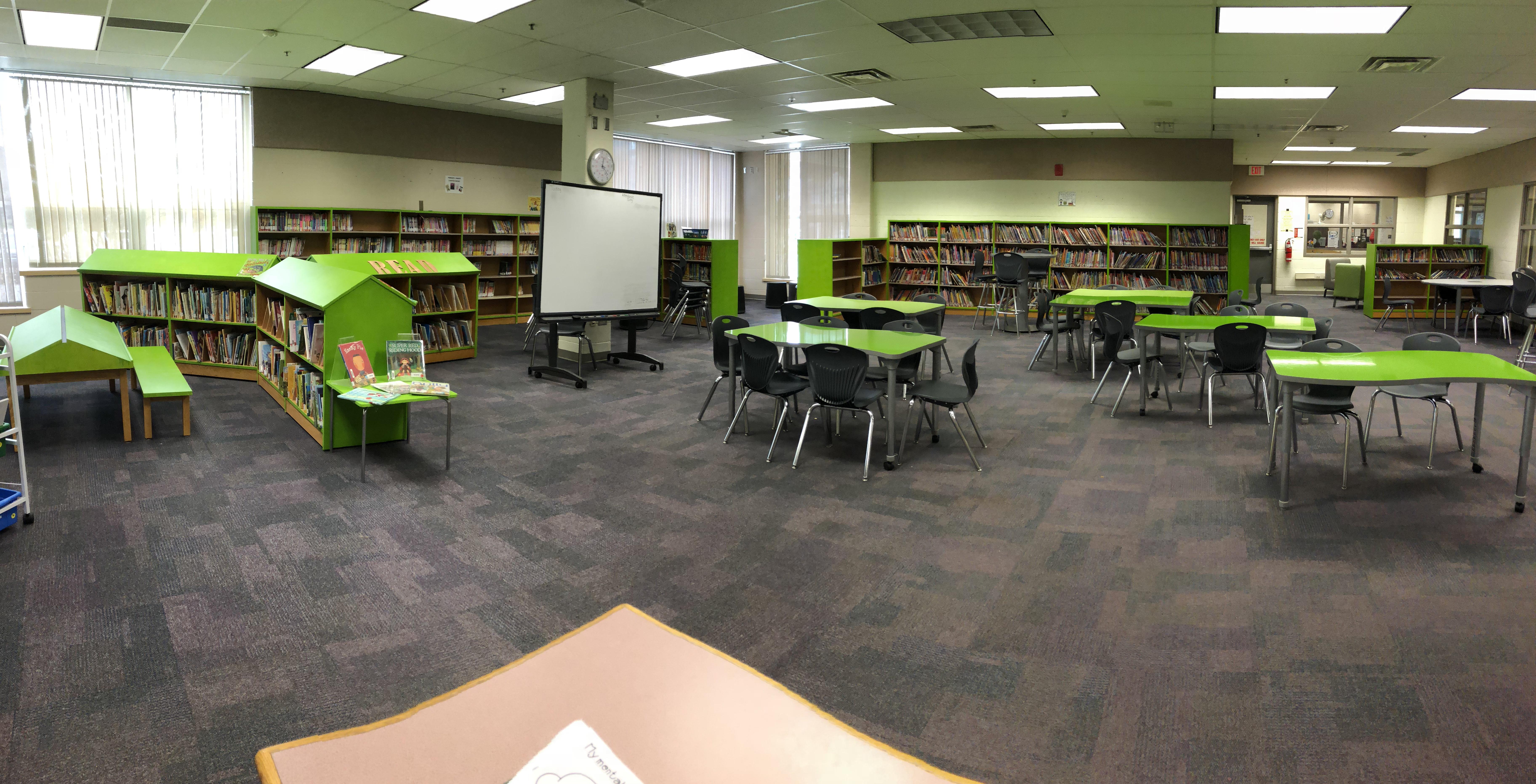 view of library, bookshelves line the wall of a large room, green tables are spread about the space with chairs at each table